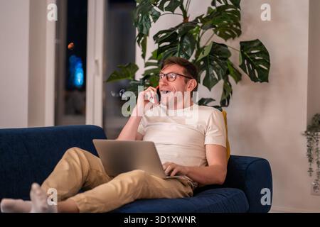Surprised middle aged man talking on mobile phone lying on couch with laptop Stock Photo