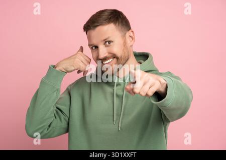 Happy bearded man pointing index finger to camera, showing phone gesture call me on pink background. Stock Photo