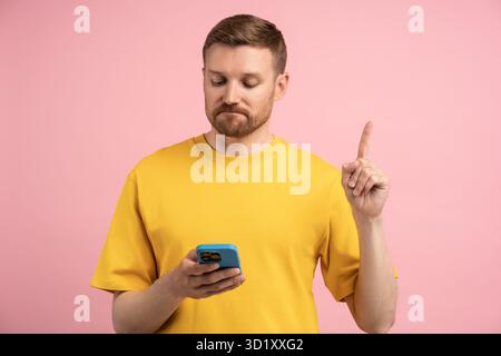 Sad upset man pointing index finger up looking in smartphone on pink background. Stock Photo