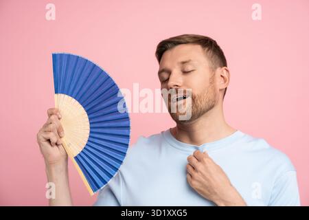 Overheat man feels relieved cooling down holds blue fan stands against pink wall studio background Stock Photo