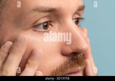 Closeup of serious bearded man looking at wrinkles under eyes touching face skin. Skincare after 40s Stock Photo