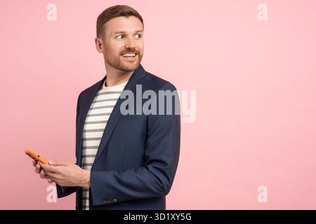 Smiling man in suit jacket with smartphone in hands looking on copy space on pink background. Stock Photo