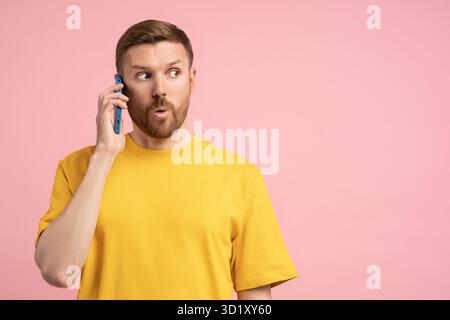 Shocked man holding cellphone surprised pointing wow eyes at copy space over pink studio background Stock Photo
