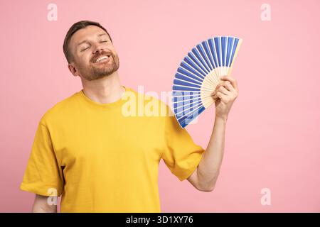 Overheated man using paper fan suffer from heat, enjoying fresh air isolated on studio pink wall Stock Photo
