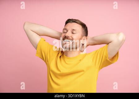 Relaxed man holding hands behind head smiling with closed eyes standing on studio pink background. Stock Photo