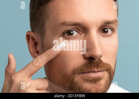 Face of serious bearded middle aged man applying face cream on cheek under eye looking at camera. Stock Photo