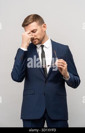 Frustrated businessman in emotional stress holds eyeglasses rubs bridge of nose on studio background Stock Photo