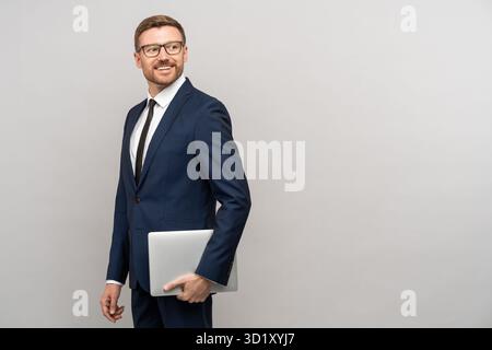 Confident happy businessman holds laptop looking on copy space on grey background. Stock Photo