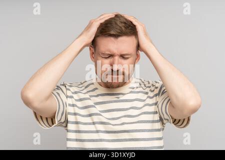 Puzzled confused bearded man holds head with closed eyes feels headache on grey background. Stock Photo