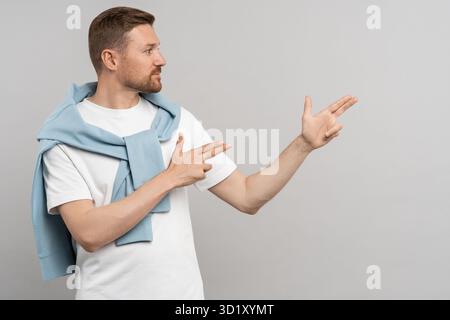 Positive young caucasian man looking and pointing fingers aside, gesturing at copy space. Stock Photo