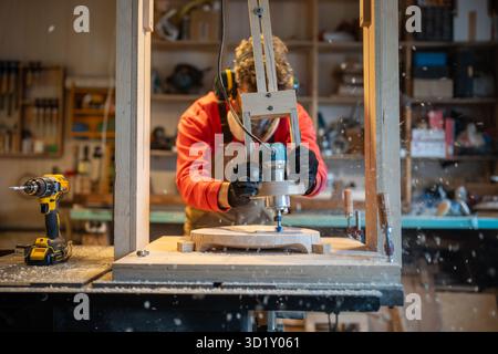 Man carpenter in noise-cancelling headphone, glasses, respirator drilling wooden plank in joinery Stock Photo