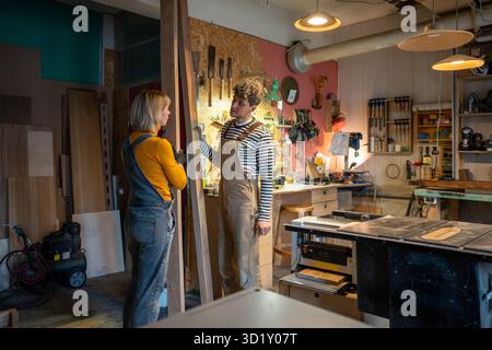 Couple of carpenters talk enthusiastically takes break from work, chatting in workshop about project Stock Photo