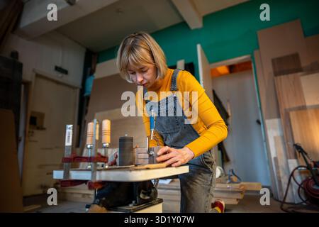 Focused woman carpenter turns wooden handmade craft products on lathe working for small business. Stock Photo