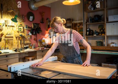 Focused woman carpenter saws board on circular saw in protective glasses observes safety precautions Stock Photo