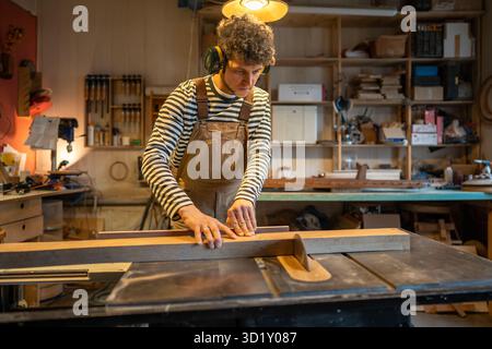 Concentrated carpenter man in protective ear muffs cuts piece of wood on circular saw in workshop. Stock Photo