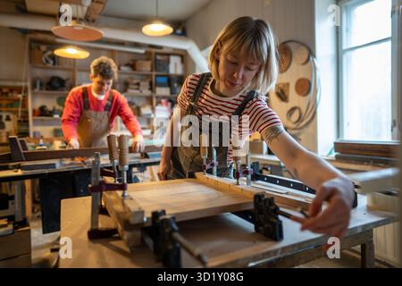 Concentrated female carpenter working on gluing wooden planks together, using bar clamps. Woodwork. Stock Photo