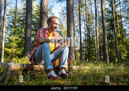 Happy interested middle aged man browsing in cellphone surfing online sitting on log in park forest. Stock Photo