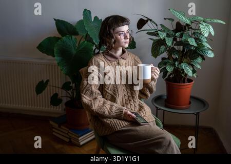 Pensive thoughtful young stylish woman in glasses holding mug looking at window at home with plants. Stock Photo