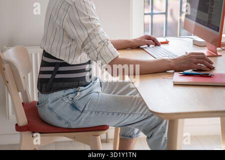Man works on computer wears back support belt on lower back to relieve stress on spine has hernia. Stock Photo