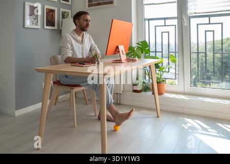 Man IT freelancer working on computer at home doing foot exercise using massage ball. Stock Photo