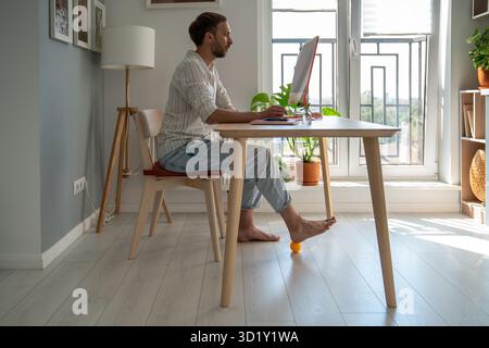 Man freelancer working on computer at home doing recovery foot exercise using massage silicone ball. Stock Photo