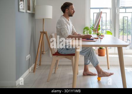 Man freelancer working on computer at home doing recovery foot exercise using massage silicone ball. Stock Photo