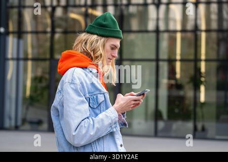 Pleased hipster guy standing outside with smartphone, looking screen typing message in social media. Stock Photo