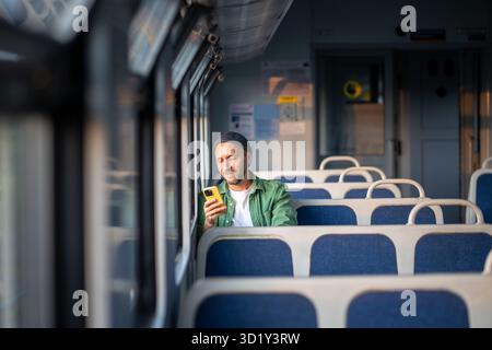 Smiling man using smartphone, scrolling social media and messaging, seated by the window in train Stock Photo