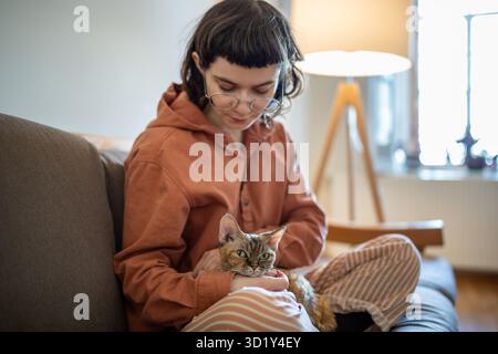 Girl stroking Devon Rex cat sitting on couch at home. Caring for pet taken from animal shelter. Stock Photo