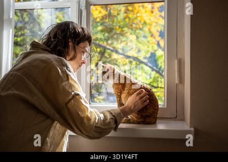 Tender pet lover stroking beloved adorable breed cat Devon Rex sitting on windowsill in sunlight Stock Photo