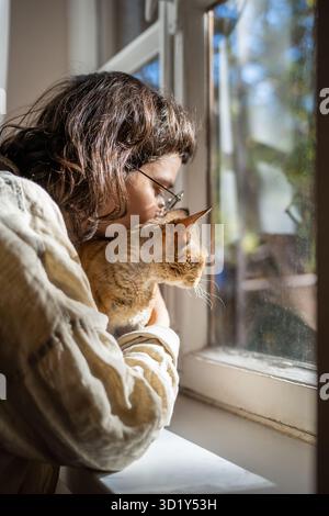 Caring pet lover cuddling beloved breed cat Devon Rex sitting on windowsill looking at window Stock Photo