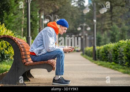 Trendy guy sitting on bench in park, waiting for girlfriend, surfing in mobile internet, messaging Stock Photo