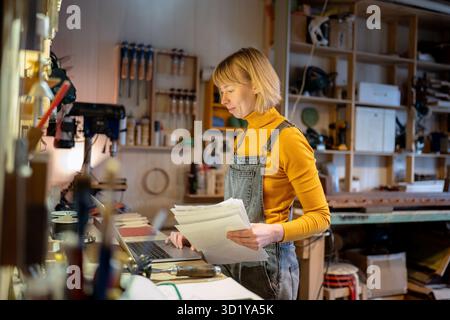 Smiling female carpenter working on laptop with client orders and personal projects for suppliers Stock Photo