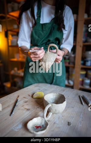 Woman potter hands refining mug handle shape with clay carving knife on table in pottery workshop Stock Photo