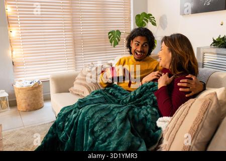 Asian couple exchanging gift box on sofa at home under dark green blanket with string lights Stock Photo