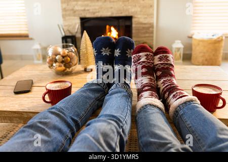 Two pairs of legs resting on coffee table in living room with red mugs ornaments fireplace Stock Photo