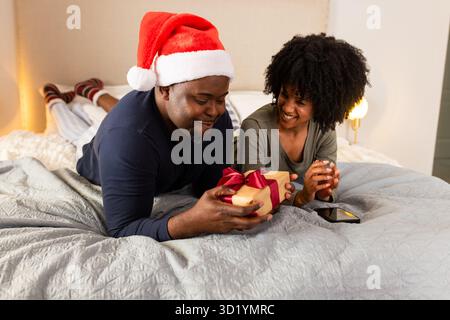 Diverse couple wearing Santa hat lying on bed exchanging gift with smartphone resting beside lamp Stock Photo