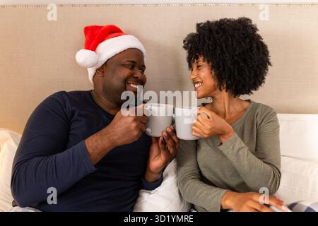 Diverse couple wearing Santa hat sitting in bedroom on bed clinking grey mugs and smiling Stock Photo