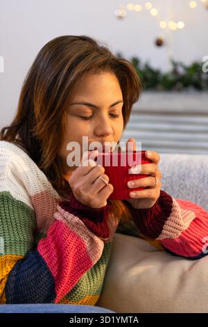 Mid-adult woman wearing striped sweater holding red mug sipping warmth on living room sofa Stock Photo