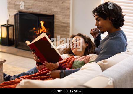 Couple reading large red-covered book on sofa with orange knit throw near stone fireplace at home Stock Photo