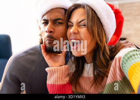 Diverse couple taking playful selfie on couch at home wearing Santa hats and festive sweaters Stock Photo