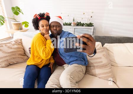 Diverse couple taking smartphone selfie while leaning on sofa in living room amid holiday garland Stock Photo