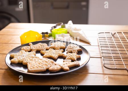 Black round plate holding icing-decorated gingerbread cookies on wooden table near colorful cutters Stock Photo