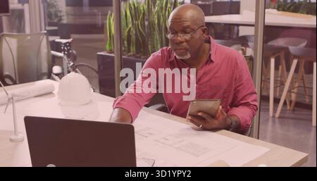Architect reviewing blueprints using laptop tablet in office wearing red shirt with white hard hat Stock Photo