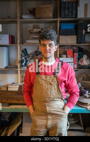 Smiling man carpenter resting in workshop looking at camera putting hands in pockets leans on table Stock Photo