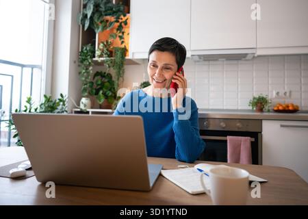 Confident mature woman smiling while talking on smartphone and working remotely on laptop at home. Stock Photo