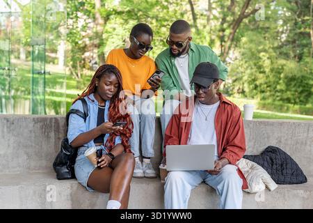 Group of interracial friends on bench work on laptops, phones. Digital native students exploring Net Stock Photo