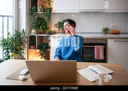 Remote boss. Smiling woman managing freelance projects with phone call and laptop in home office Stock Photo