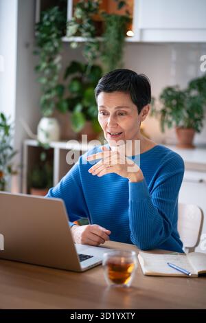 Focused mature woman gestures as explains tasks on video call at home, stting with tea and notes Stock Photo