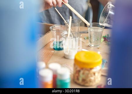 Hands pouring resin into clear cups for crafting project on worktable with paint and materials. Stock Photo